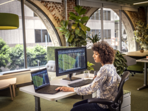 woman sitting at a desk in an office with a laptop and monitor doing computer drafting.